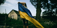 Ein junger Mann steht in einem Vorgarten eines kleinen Dorfes in der Region Charkiw und hisst eine Ukraineflagge